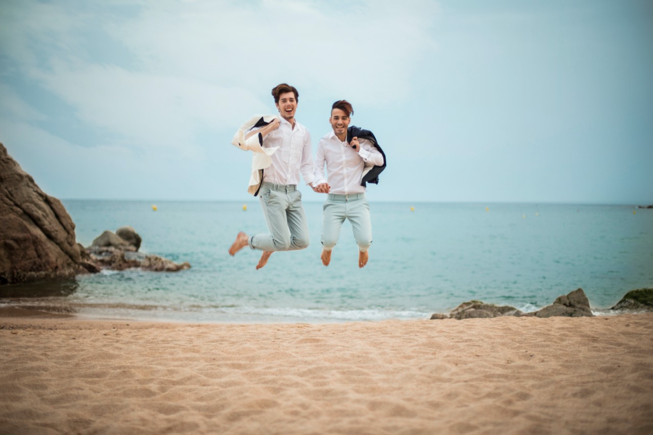 Two men in light wedding wear jumping on the beach
